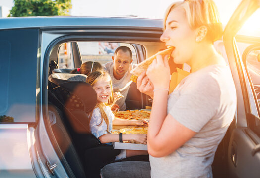 Positive smiling girl in child car seat while family car trip brake stop eating just cooked Italian pizza. Happy family moments, childhood, fast food eating or auto journey lunch break concept image.