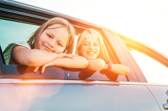 Two happy sisters looking out open car window during auto trip. Cute girls are smiling, laughing during road jorney. Family values, traveling concept side car view photo.