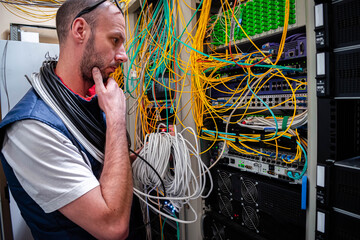 A man with a large skein of wires is working in a server room. A technician is thinking about how to switch the Internet cables in a data center.