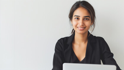 Indian female assistant, working on a laptop, wearing black corporate clothes, white wall background 