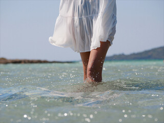 A girl in a white dress walks into the sea. Legs close up.