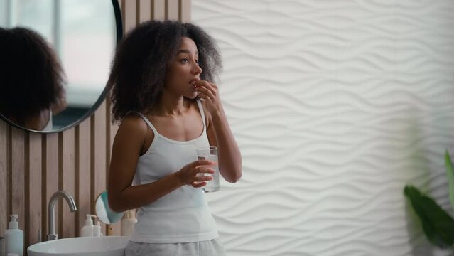 Healthy African American beautiful woman sick ill girl female taking pill with glass of water drinking remedy drug vitamins supplements at morning bathroom daily medication for beauty skin hair care
