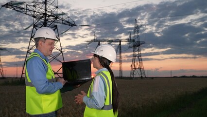 Man electrician talks to lady trainee showing information project data on laptop near power...