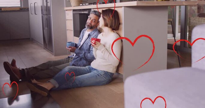 Animation of hearts over diverse couple drinking coffe and sitting on floor in kitchen at home