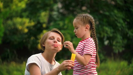 Loving mother with cheerful daughter blows bubbles in park among trees on vacation. Mother waits till preschooler girl blows bubbles in meadow on weekend. Mother and child spend time blowing bubbles