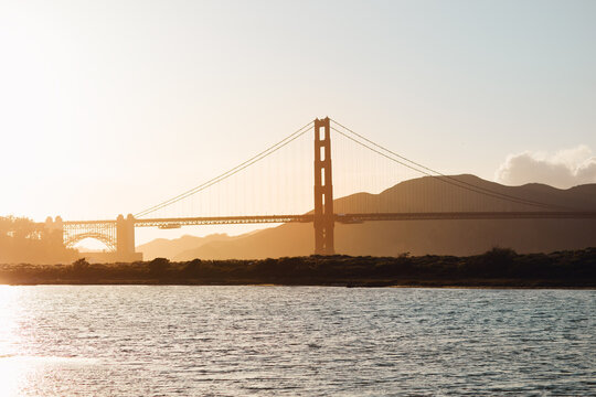 The setting sun casts a golden silhouette of the Golden Gate Bridge, creating a serene scene over the bay with hazy mountain backdrops