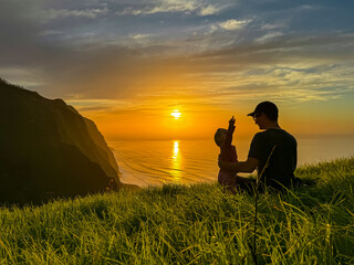 Father playing with toddler on meadow with breathtaking sunset at viewing point Miradouro do Ponta da Ladeira, Madeira island, Portugal, Europe. Panoramic view of majestic coastline Atlantic Ocean