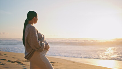 Pregnant woman looking sunset standing on sandy beach. Girl caressing belly