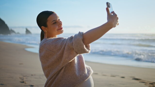 Pregnant woman making selfie standing sandy seashore caressing belly close up.