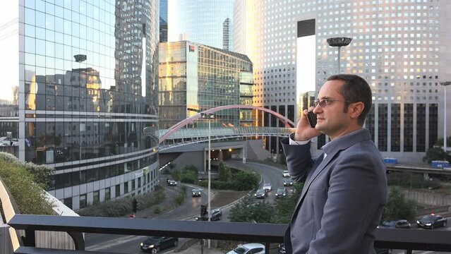Businessman talks on phone on balcony of his office in front of modern skyscrapers