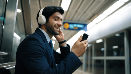 Smart business man listening music by headphone while waiting train at train station with blurring background. Skilled project manager enjoy listen relax sound while holding mobile phone. Exultant. - obrazy, fototapety, plakaty