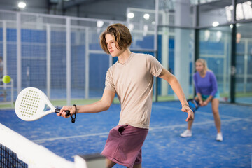 Teenage boy serving ball while playing padel in court during training.