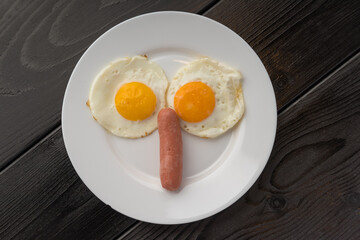 Two fried eggs looking like a face with a sausage for a smile on a white plate, dark wooden surface.