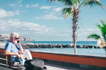 Fototapeta na wymiar Senior bearded man holding smartphone sitting outdoors on bench face the sea enjoying sunny day, travel, vacation, retirement lifestyle