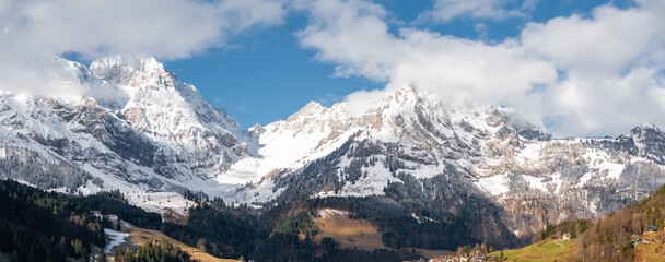 Panoramic view of Engelberg, Switzerland, showing snowcapped mountains, evergreen trees, and a valley with alpine buildings under a partly cloudy sky, highlighting nature's grandeur.
