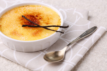 Delicious creme brulee in bowl, vanilla pods and spoon on light textured table, closeup