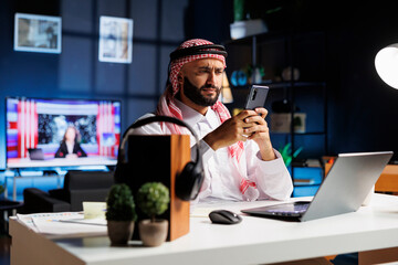 Professional Arab man works in an office, using a computer and smartphone for online research and...