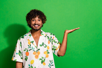 Photo of excited funky latin guy dressed print shirt smiling showing arm empty space isolated green...