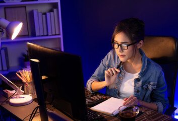 Thinking young beautiful Asian creative woman looking on pc with taking note on paper for memo in...