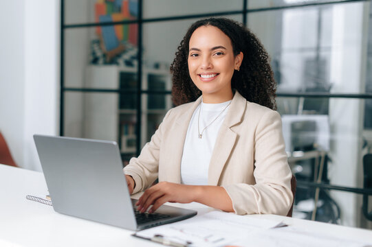 Photo of a beautiful curly haired brazilian or hispanic business lady, project manager, in a suit, sitting in a creative office space while working in a laptop, looks at camera, smile friendly