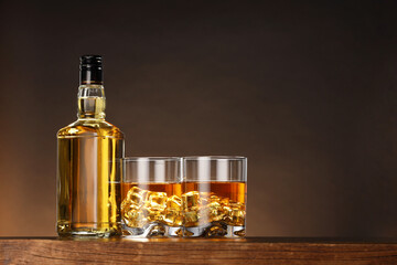 Whiskey with ice cubes in glasses and bottle on wooden table against brown background, space for text