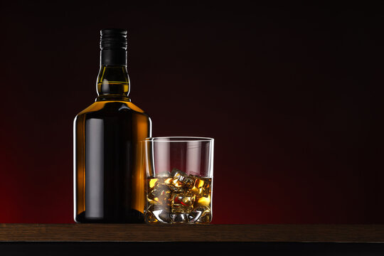 Whiskey with ice cubes in glass and bottle on wooden table, space for text