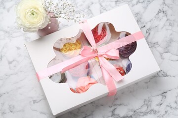 Different colorful cupcakes in box and flowers on white marble table, above view
