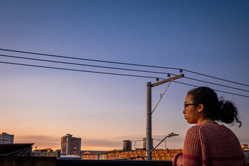 Woman standing and wearing pink sweater on terrace in latin city with an electric tower at sunset