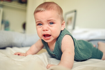Close up portrait of little funny cute blonde infant boy child toddler with blue eyes in green linen bodysuit crying indoors at nursery room. Childish tantrum. Difficulty problems putting baby to bed