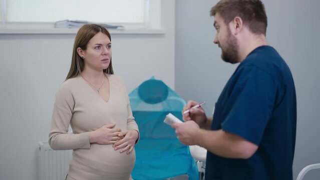 A doctor gives recommendations to a pregnant woman while standing near a gynecological chair. The woman holds her hands on her belly and nods her head in agreement with the doctor