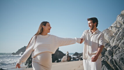 Laughing pregnant spouses walking sunny rocky beach feeling happiness close up.