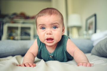 Close up portrait of little funny cute blonde infant boy child toddler with blue eyes in green linen bodysuit crying indoors at nursery room. Childish tantrum. Difficulty problems putting baby to bed