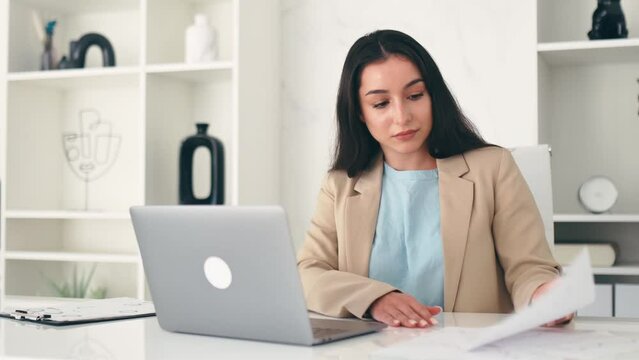 Concentrated serious indian or arabian business woman, financial manager, sitting at workplace in the office, using laptop, doing paperwork, focused studying documents, create business plan