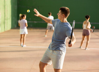 Positive motivated young frontenis player swinging string racquet to hit ball on outdoor walled court on sunny summer day. Sport and active lifestyle concept