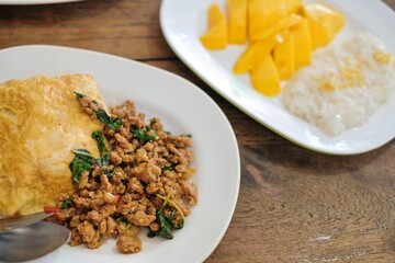 A plate of pad kra pao (holy basil pork mince stir-fry) with omelette and mixed grain rice, and a dessert of fresh mango with sticky rice at a street food stall in Phra Nakhon, Bangkok, Thailand - obrazy, fototapety, plakaty
