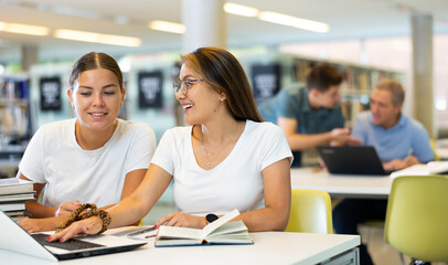 Two positive ladies wearing casual clothes behave loudly while studying in the library and using computer