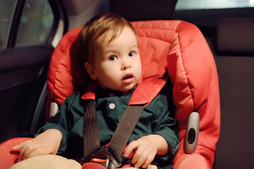 Cute toddler boy is in car seat. Portrait of pretty little child during family road trip. Kid scared by motion sickness or riding in vehicle at night time. Safety transportation of baby by car. - obrazy, fototapety, plakaty