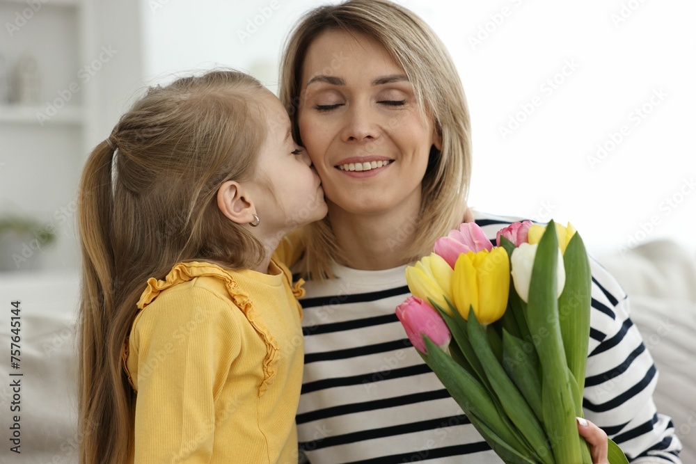 Wall mural Little daughter kissing and congratulating her mom with Mother`s Day at home. Woman holding bouquet of beautiful tulips