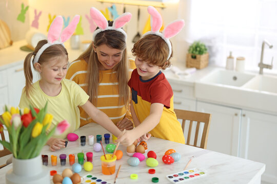 Easter celebration. Mother and her cute children with bunny ears painting eggs at white marble table in kitchen