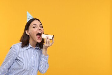 Happy young woman in party hat eating cheesecake on yellow background