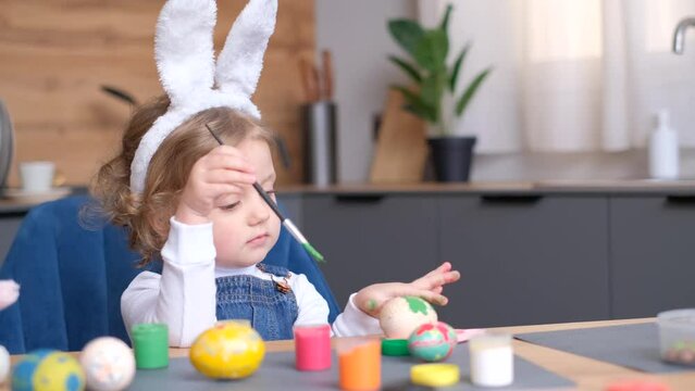 A little girl in bunny ears painting Easter eggs at a table