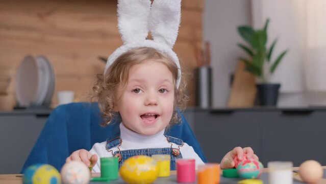 Toddler with bunny ears decorates Easter eggs at table, smiling happily