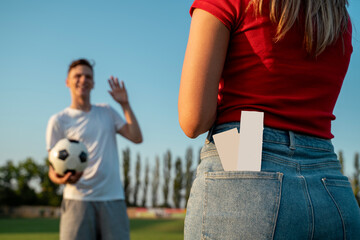 Closeup shot, girl with two blank tickets in back pocket and her friend with soccer ball, going on soccer match concept 