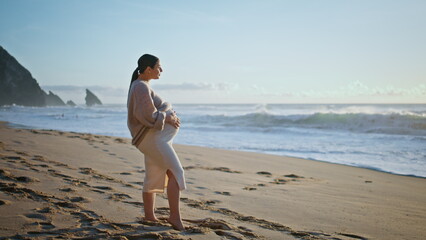 Beautiful pregnant woman posing sandy beach contemplating foamy ocean waves.