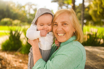 Beautiful happy smiling senior elderly woman holding on hands cute little baby boy. Grandmother and grandson having fun time together at tropical summer day in park. Multigenerational family concept