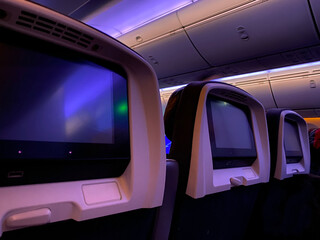 Interior of an airplane with digital monitor to watch movies during the flights. View of the seats, screen, luggage racks and front of the aircraft. Dim lighting for a night flight.