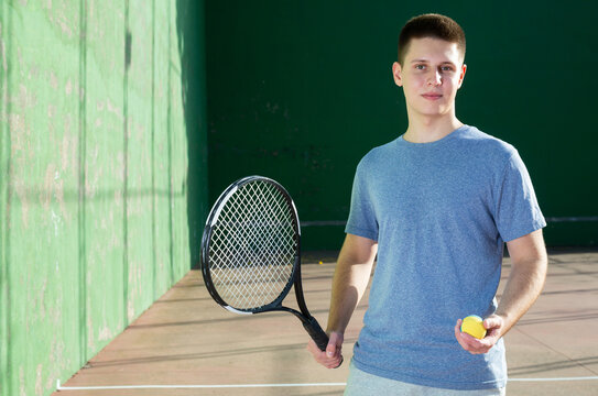 Portrait of positive young man frontenis player standing on outdoor fronton, holding racket and rubber ball and looking at camera.