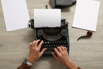 Mature man with paper sheets and smoking pipe typing on vintage typewriter at beige wooden table