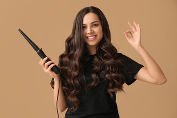 Beautiful young woman with curling iron showing OK on beige background