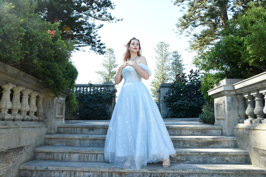 Close up  portrait of beautiful female model wearing blue fantasy ballgown, like a fairytale elf princess.  Elegant pose, hands reaching out, on the balcony terrace of a romantic castle location.
